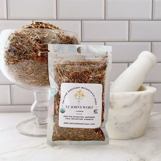 A clear plastic bag of organic St John's Wort Herb sits in front of a glass bowl filled with loose herbs, renowned for their metaphysical properties. A white mortar and pestle is positioned to the right. Both the bowl and bag are placed on a white tiled surface against a white brick background.