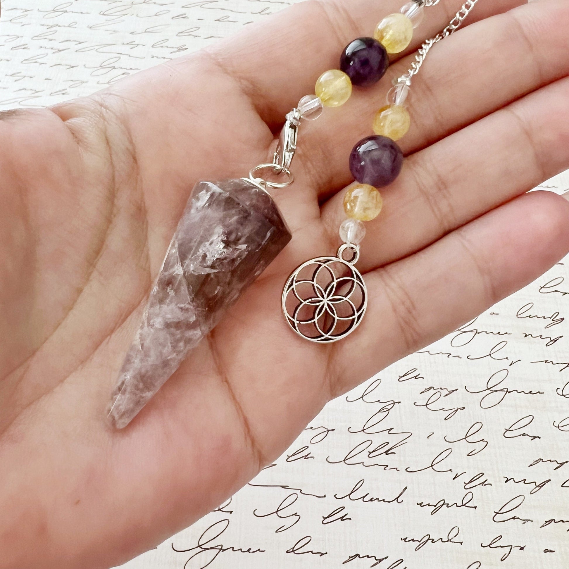 A Super Seven Hexagonal Pendulum with Seed of Life Charm, an exquisite energy healing tool with a chain adorned with clear, yellow, and purple beads, lies on a notebook filled with cursive writing. The chain features a silver pendant showcasing an intricate geometric design.