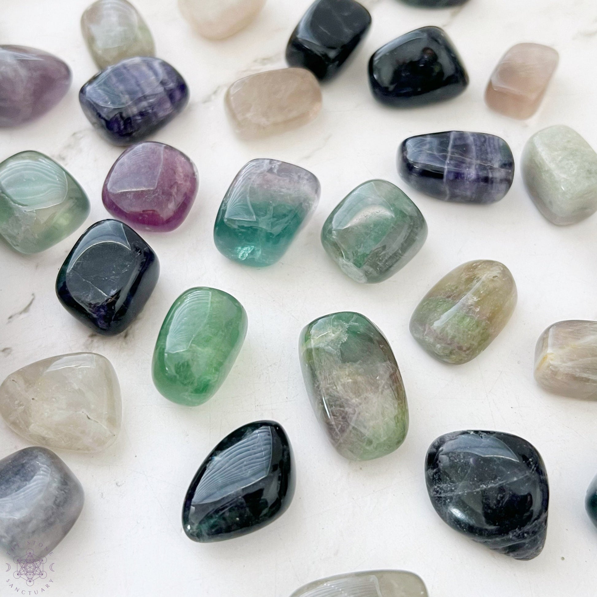 A wooden bowl filled with a collection of polished Rainbow Fluorite Tumbled Stones in various colors. The stones range in hues from green and blue to purple and clear, with smooth, rounded shapes. The bowl rests on a white background, showcasing these healing crystals beautifully.