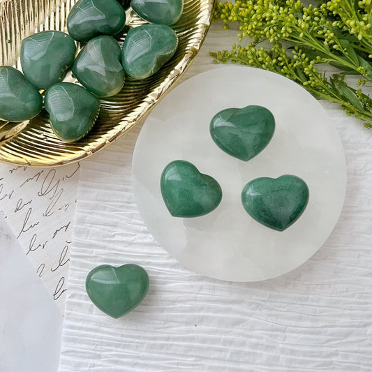 A collection of green heart-shaped 1" Mini Green Aventurine Pocket Hearts is displayed on a white surface. Some stones are placed on a circular white tray, while others rest on a gold-colored leaf-shaped tray. Green foliage partially frames the upper right corner of the image, manifesting prosperity and abundance.