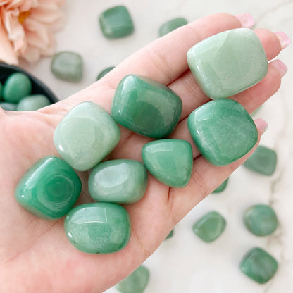 A wooden bowl filled with smooth, polished Green Aventurine Tumbled Stones, known as the "Stone of Opportunity." The stones are uniform in size with a glossy finish, enhancing their various shades of green. Often associated with the Heart Chakra, the bowl is placed against a plain white background.