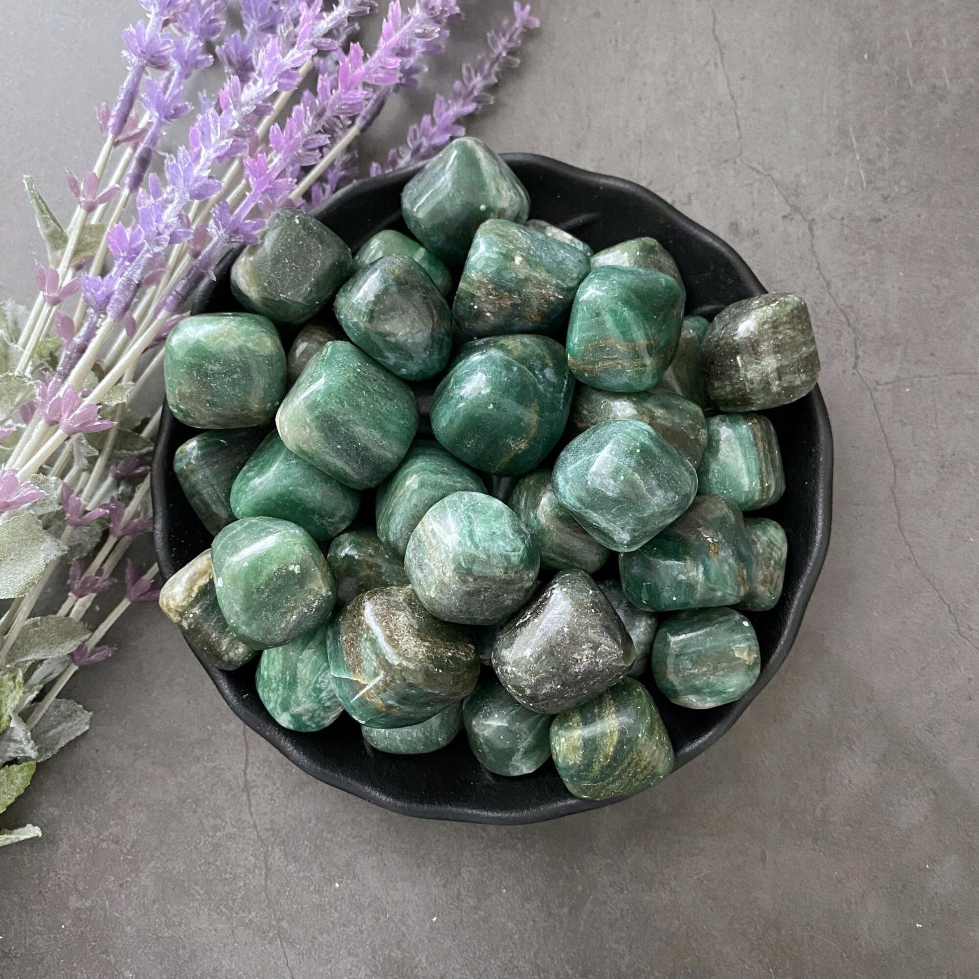 A black bowl filled with Emerald in Matrix Tumbled Stones, resembling emerald crystals, sits on a gray surface. Sprigs of purple lavender are positioned on the left side of the bowl, adding a contrasting element to the arrangement and enhancing its connection to the heart chakra.