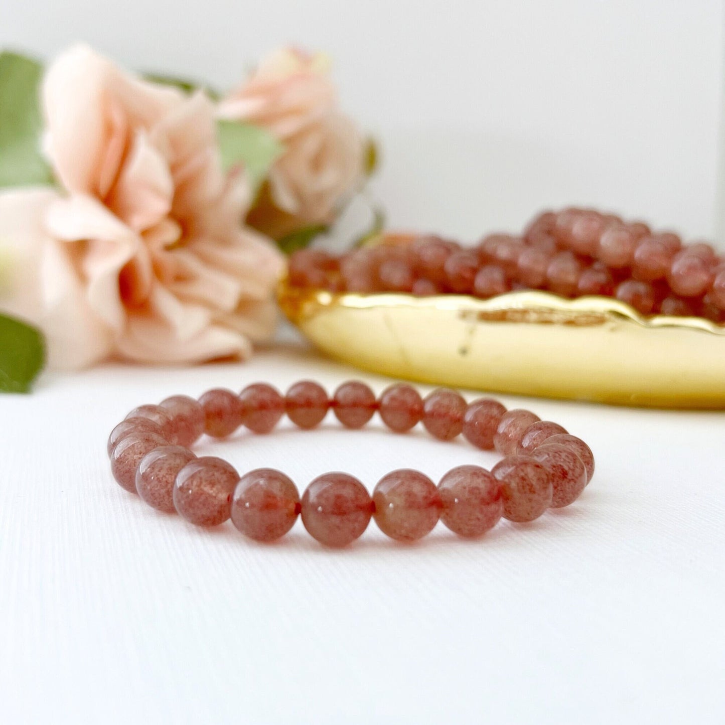 An 8mm Strawberry Quartz Beaded Bracelet made of spherical, peach-colored beads rests on a white surface. In the background, there are peach-colored roses slightly blurred, and a golden dish filled with similar beads. The overall setting is elegant and softly lit.