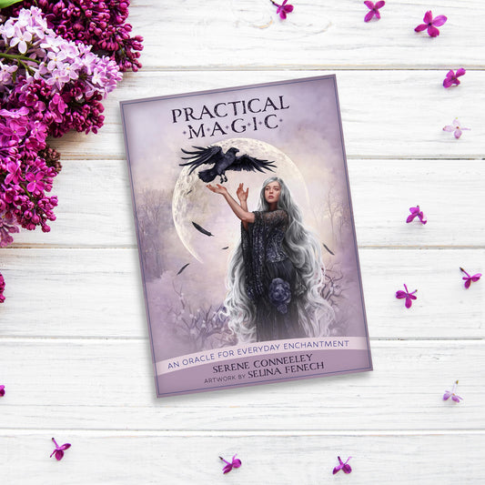 A book titled "Practical Magic: An Oracle for Everyday Enchantment" by Serene Conneeley, with artwork by Selina Fenech, lies on a white wooden surface surrounded by lilac flowers. Alongside the Practical Magic: An Oracle Deck for Everyday Enchantment, the cover features a woman with long, flowing hair and a raven set against a mystical backdrop.