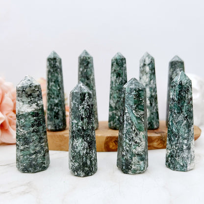 A set of eight 3-4" Emerald Points arranged in a semi-circle on a white surface. In the background, there are pink flowers and a white pot with leaves and the text "Give Kindness daily" printed on it, creating an energy generator for the heart chakra.