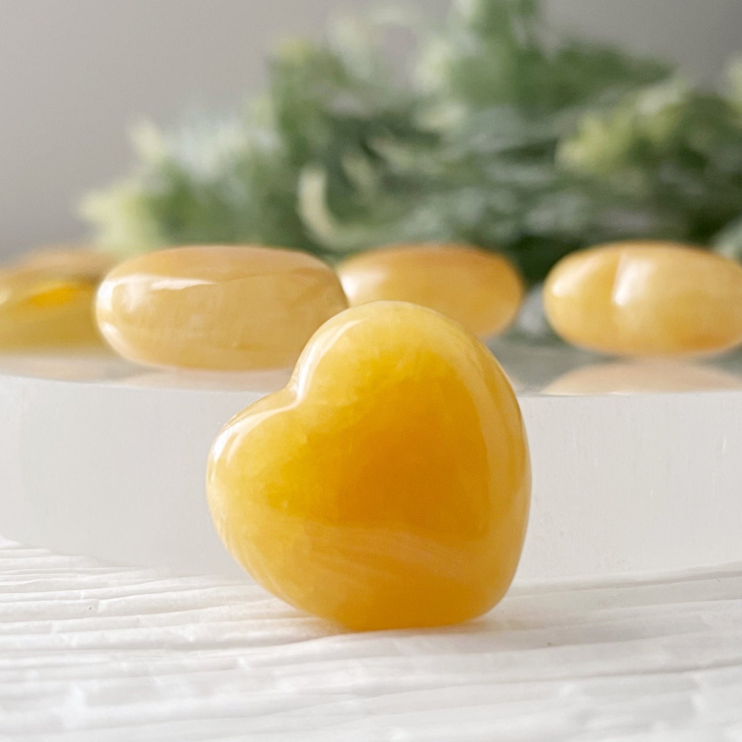 A collection of polished, 1" Mini Yellow Calcite Heart stones displayed on a round white surface, with more stones in a gold dish nearby. The background features a textured white fabric and a hint of green foliage in the upper right corner, perfect for crystal healing and balancing the Solar Plexus Chakra.