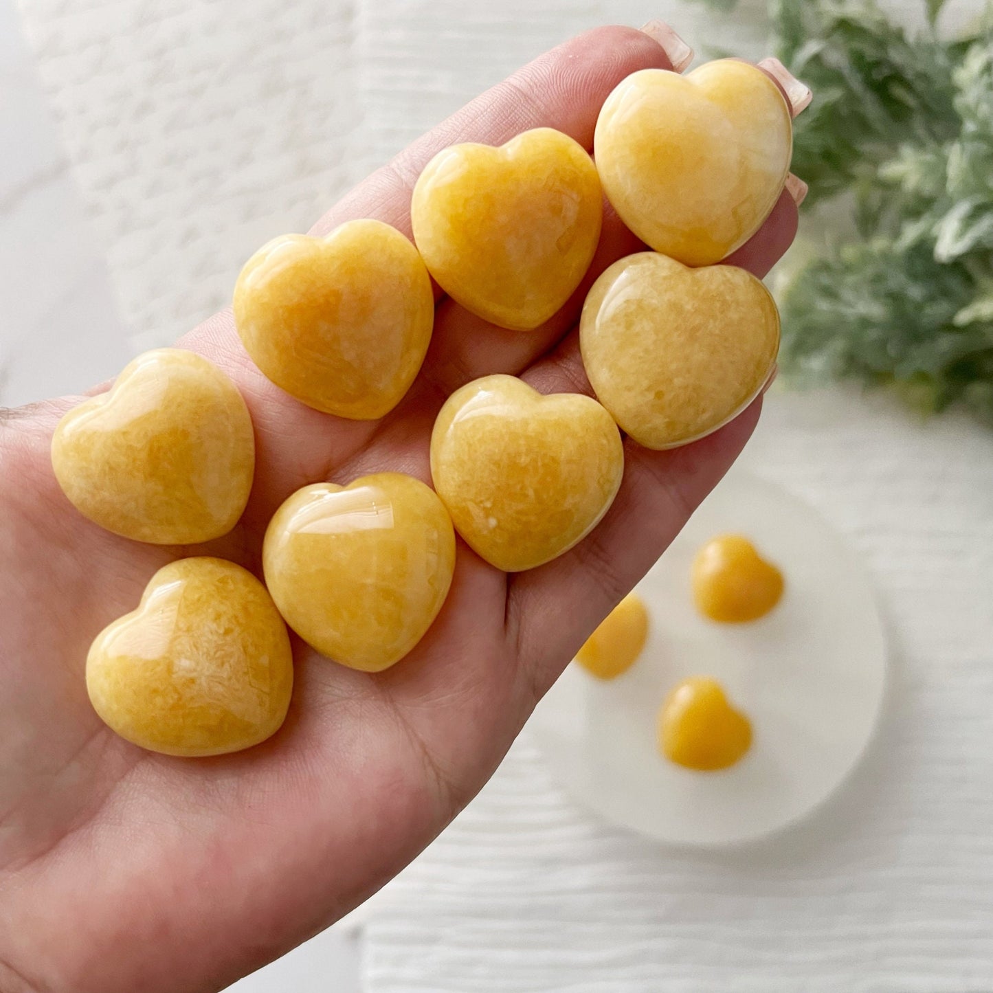 A collection of polished, 1" Mini Yellow Calcite Heart stones displayed on a round white surface, with more stones in a gold dish nearby. The background features a textured white fabric and a hint of green foliage in the upper right corner, perfect for crystal healing and balancing the Solar Plexus Chakra.