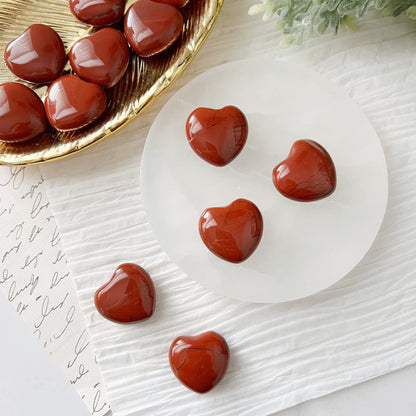 A decorative setup featuring red heart-shaped confections and 1" Mini Red Jasper Hearts. Several hearts are placed on a round white marble platter, with more in a gold dish nearby. The arrangement is on a white crumpled sheet, accented with greenery and a handwritten script underneath.