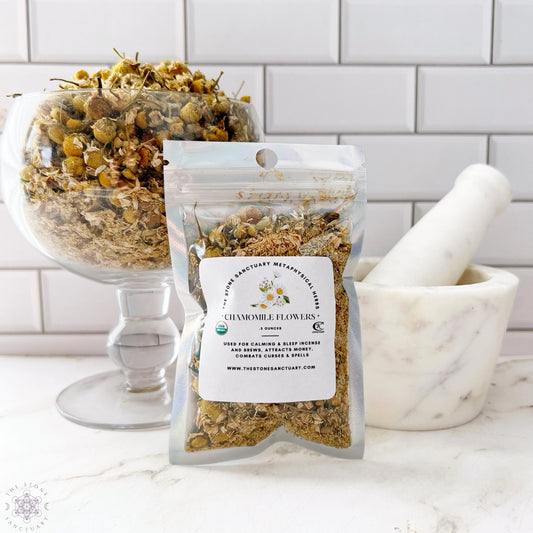 A sealed pack of Chamomile Flowers is displayed upright next to a clear glass bowl filled with loose chamomile flowers and a white marble mortar and pestle. The background features a tiled wall with a subtle pattern.