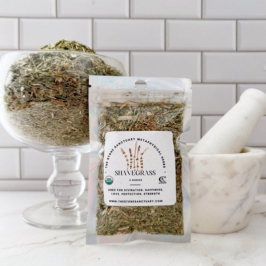 A white kitchen counter holds a large glass bowl filled with dried herbs, a white mortar and pestle, and a small clear pouch labeled "Shavegrass Herb." The label mentions metaphysical uses for divination, happiness, love, protection, and strength. A tile backsplash is visible behind.