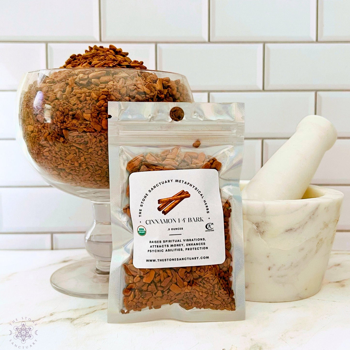 A small silver bag labeled "Cinnamon Chips" with information on the front stands next to a white mortar and pestle. Behind them is a large glass bowl filled with cinnamon bark pieces, perfect for enhancing your culinary endeavors. The background features a white tiled wall.