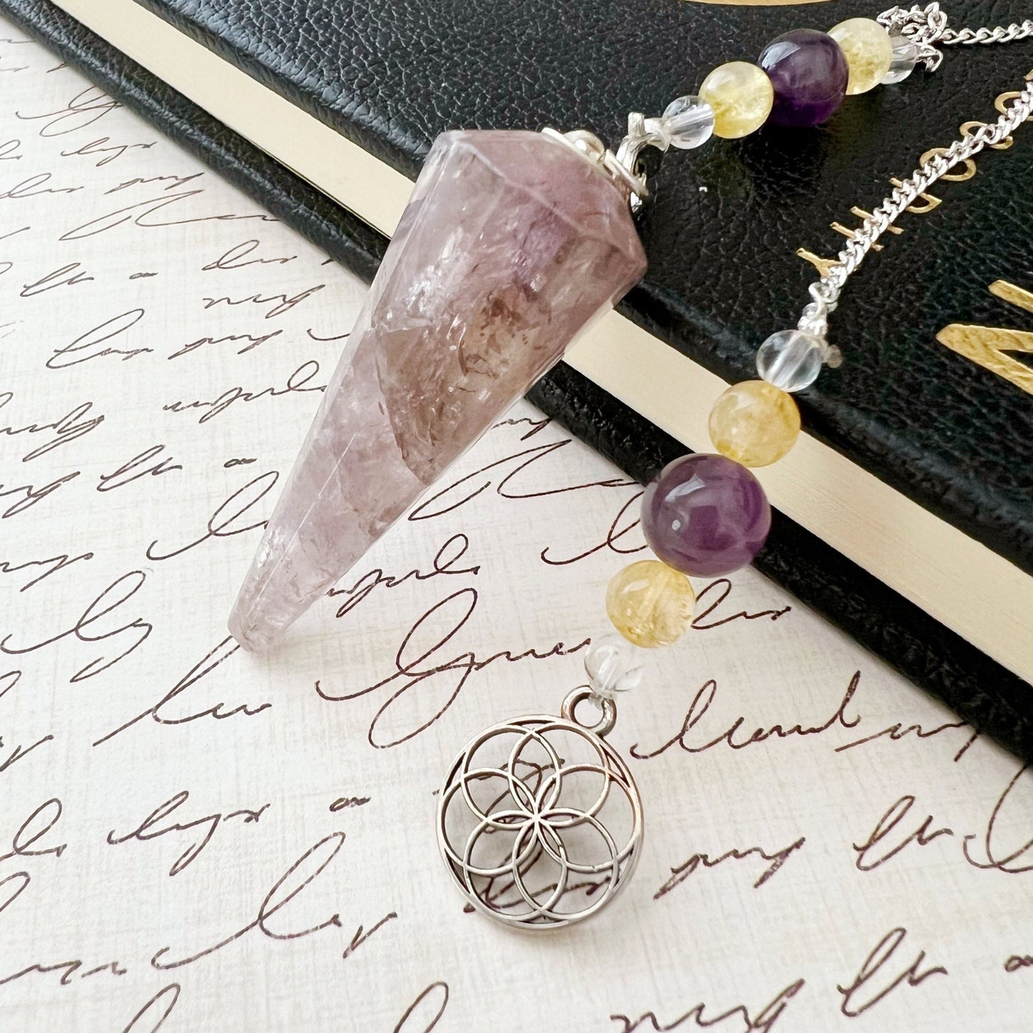 A Super Seven Hexagonal Pendulum with Seed of Life Charm, an exquisite energy healing tool with a chain adorned with clear, yellow, and purple beads, lies on a notebook filled with cursive writing. The chain features a silver pendant showcasing an intricate geometric design.