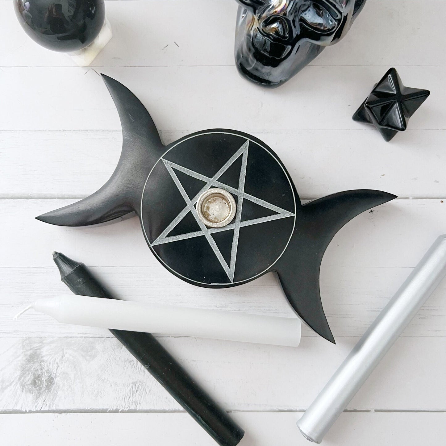 A 6" Black Soapstone Pentacle Candle Holder shaped like a crescent moon is centered on a white wooden surface. Surrounding it are chime spell candles, a black skull, an obsidian crystal, and other decorative items. The scene has a mystical, ritualistic aesthetic.