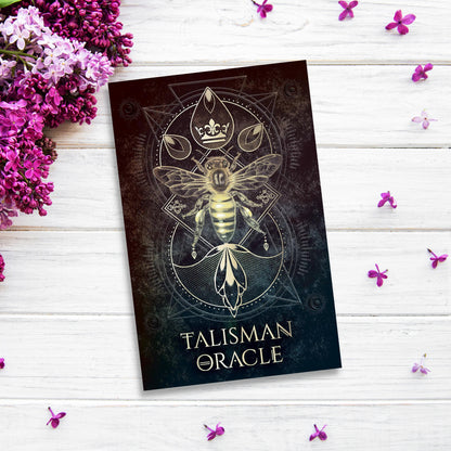 A card with a decorative bee design titled "Talisman Oracle" from the black gilded-edged Talisman Oracle is placed on a wooden surface. Purple flowers and scattered petals surround the card, creating an elegant and mystical ambiance.