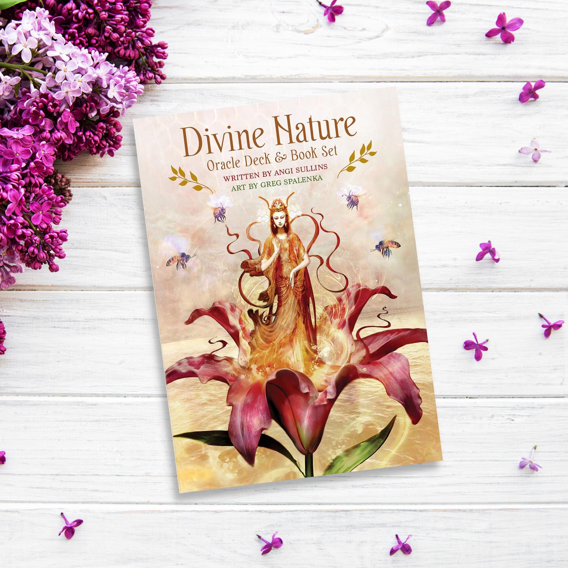 A Divine Nature Oracle Card Deck & Book Set lies on a white wooden surface with lilac flowers and small purple petals scattered around. The cover features an ethereal figure standing amidst large flowers and bees, with artwork by Greg Spalenka. This enchanting set includes gold gilded-edged cards and a 100-page guidebook.