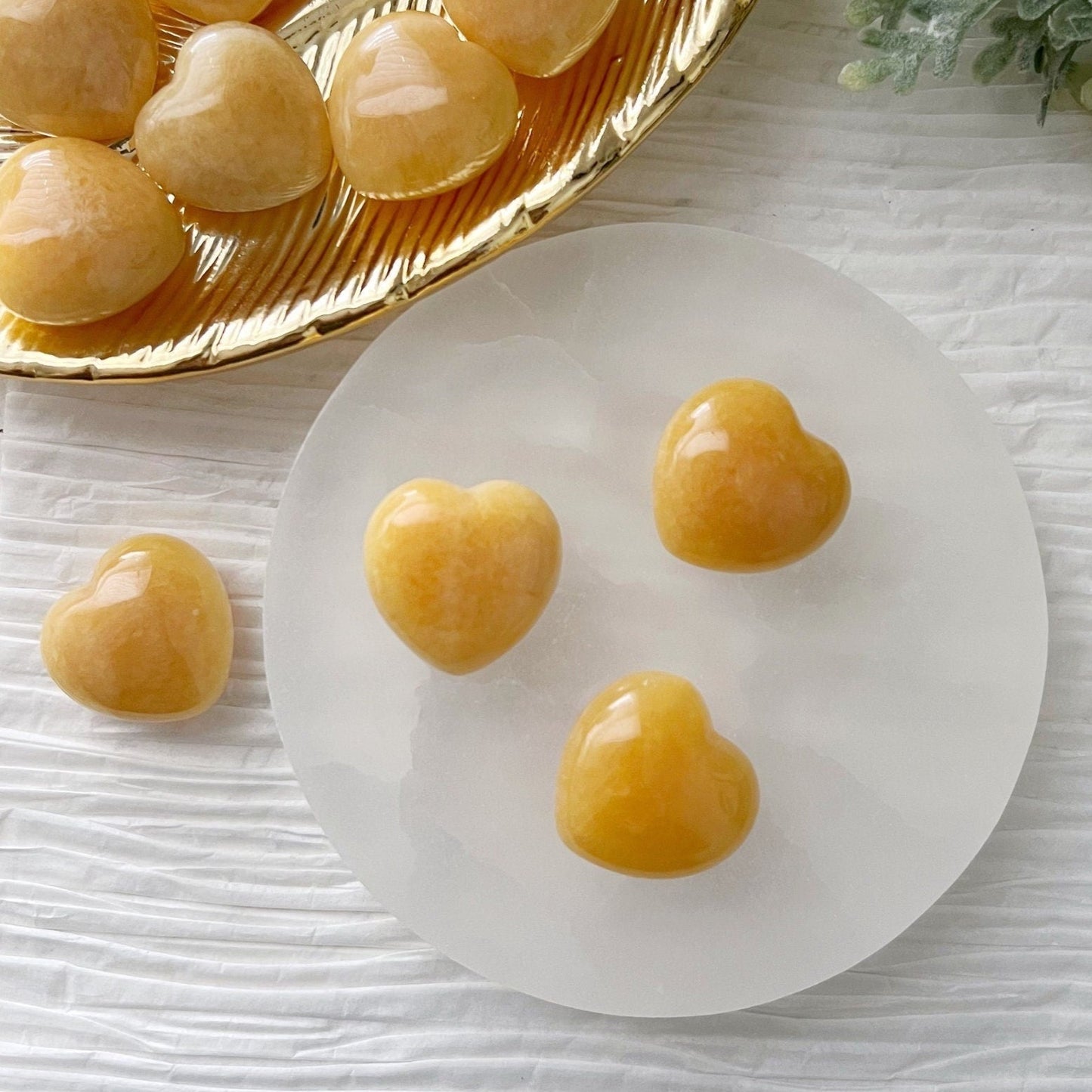 A collection of polished, 1" Mini Yellow Calcite Heart stones displayed on a round white surface, with more stones in a gold dish nearby. The background features a textured white fabric and a hint of green foliage in the upper right corner, perfect for crystal healing and balancing the Solar Plexus Chakra.