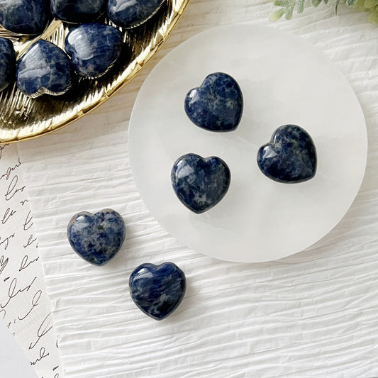 A round white plate holds five polished, heart-shaped stones, including a radiant 1" Mini Sodalite Heart. In the top left corner, a gold-rimmed dish is partially visible, also containing similarly shaped treasures. The background features white textured surfaces and a piece of paper with cursive writing, fostering inner peace.