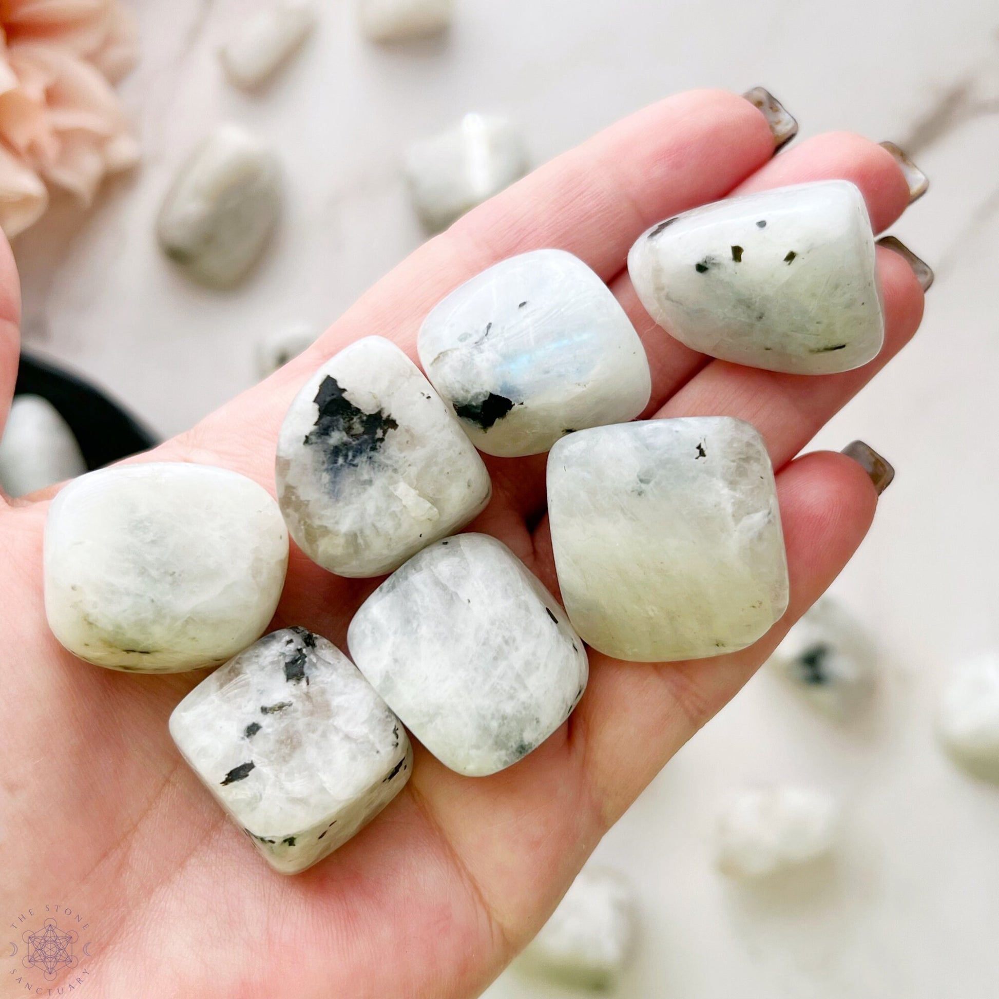 A wooden bowl filled with polished Rainbow Moonstone Tumbled Stones, varying in size and displaying a range of milky white to slightly translucent hues with occasional black specks, offers a touch of psychic protection. The bowl is set against a plain white background.