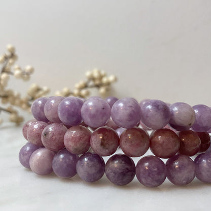 A stack of round, polished 8mm Lepidolite Beaded Bracelets in varying shades of purple and pink is displayed on a white surface. In the blurred background are small white berries on delicate branches, enhancing the calming Usui Reiki energy that these mood stabilizers offer.