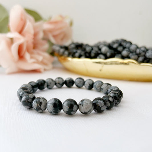 A delicate 8mm Snowflake Obsidian Beaded Bracelet made of round black and gray speckled beads is displayed against a white background. Nearby, there is a soft-focus flower and a golden dish filled with similar beads, radiating healing energy.