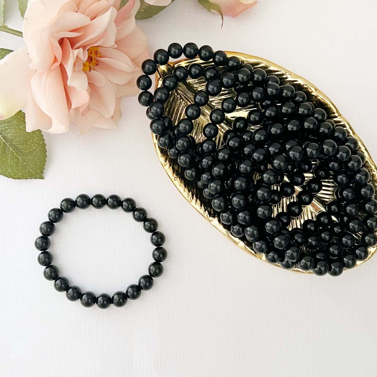 A polished 8mm Black Obsidian Beaded Bracelet is displayed on a white surface, with a bunch of pink roses in the background. Additional black beads, known for their protective qualities, are scattered on a gold-colored tray behind the bracelet, contributing to the elegant and serene composition.