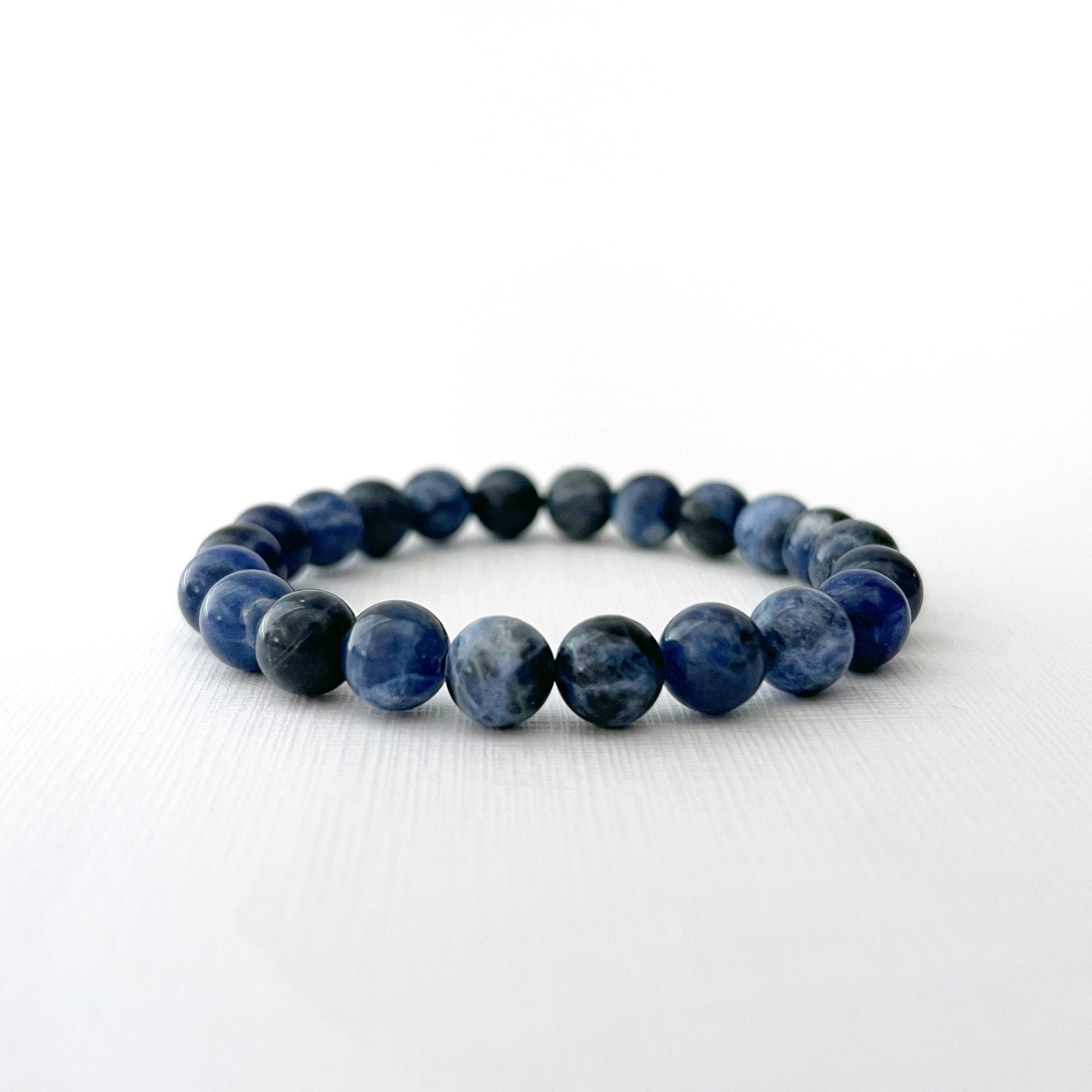 A 8mm Sodalite Beaded Bracelet lies on a white surface. In the background, there are soft pink roses and a bowl filled with blueberries, both slightly out of focus. The setting gives a serene and elegant feel, emphasizing the bracelet's metaphysical properties.