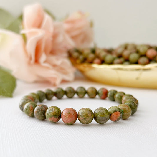 An 8mm Unakite Beaded Bracelet made of green and pink stones is displayed on a white surface. In the background, there is a blurred image of pink flowers and a golden dish filled with similar stones. The overall setting, emphasizing emotional healing, conveys a natural and elegant aesthetic.