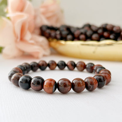 An 8mm Red Tiger Eye Beaded Bracelet featuring alternating dark brown and black polished round stones with Red Tiger Eye accents is laid out on a white surface. In the background, soft pink flowers and more beaded jewelry in a gold dish can be seen.