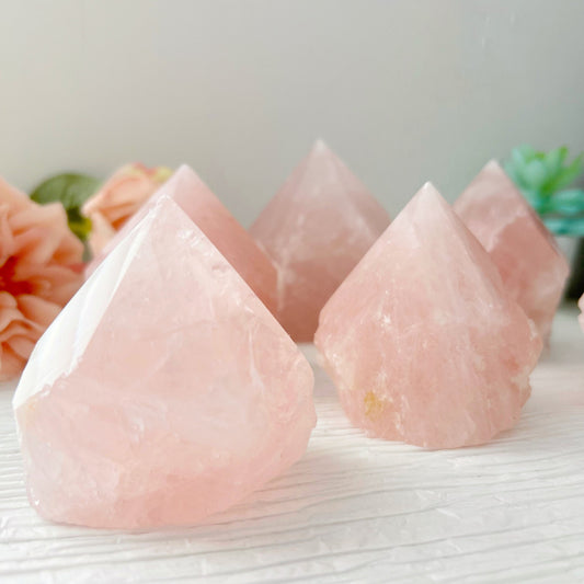 Five 2-4" Rose Quartz Top Polished Points are displayed on a light-colored surface. In the background, there are soft-focus pink flowers and green succulents, creating a serene and natural setting that embodies the essence of crystal healing.