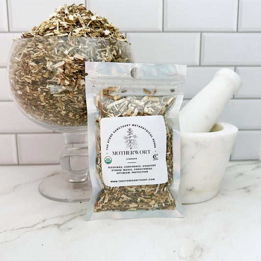 A clear plastic bag labeled "Motherwort Herb," filled with dried herb pieces, sits on a white marble surface in front of a large glass bowl filled with more dried herbs. A white mortar and pestle are positioned to the right. The setup looks perfect for magical rituals or spell casting against the backdrop of a white tiled wall.