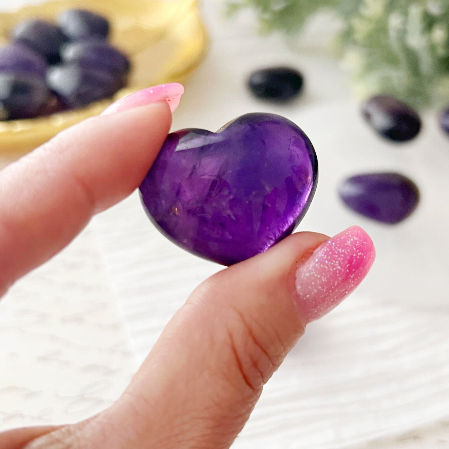 A group of polished 1" Mini Amethyst Crystal Pocket Hearts are displayed on a white textured surface. Four hearts are arranged on a white circular dish, while several more rest on a gold tray, symbolizing emotional harmony. Greenery decorates the background, adding a touch of natural elegance.