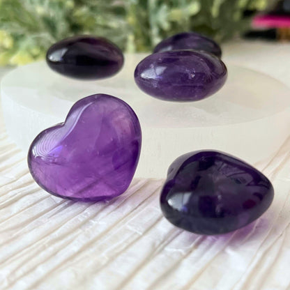 A group of polished 1" Mini Amethyst Crystal Pocket Hearts are displayed on a white textured surface. Four hearts are arranged on a white circular dish, while several more rest on a gold tray, symbolizing emotional harmony. Greenery decorates the background, adding a touch of natural elegance.