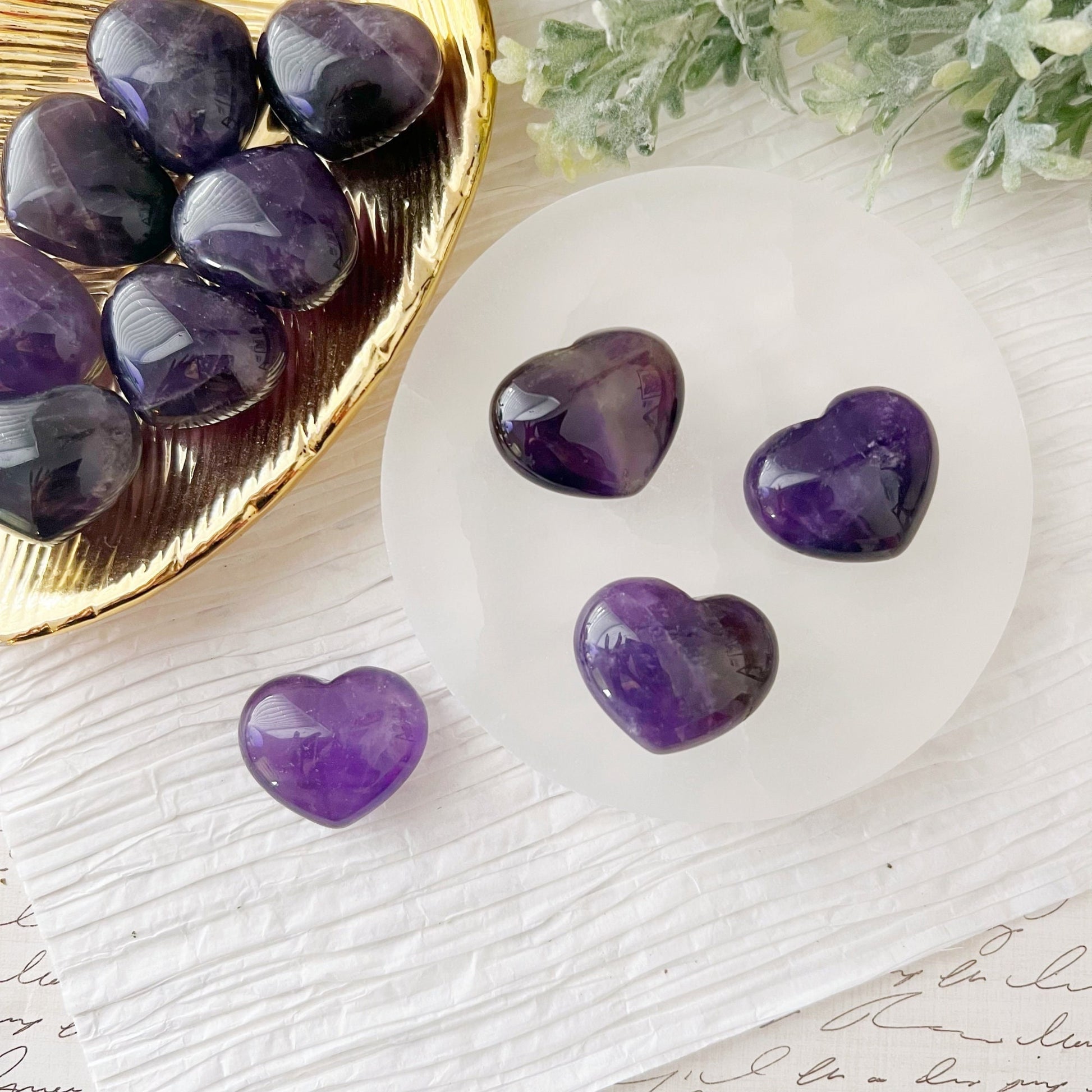 A group of polished 1" Mini Amethyst Crystal Pocket Hearts are displayed on a white textured surface. Four hearts are arranged on a white circular dish, while several more rest on a gold tray, symbolizing emotional harmony. Greenery decorates the background, adding a touch of natural elegance.