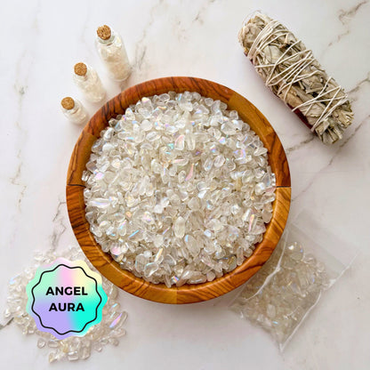 A wooden bowl filled with shiny, iridescent Angel Aura Quartz Crystal Chips labeled "Angel Aura" sits on a white marble surface. Nearby are three small vials of similar high vibe crystals and a bundle of white sage. A plastic bag with a few crystal chips spills slightly.