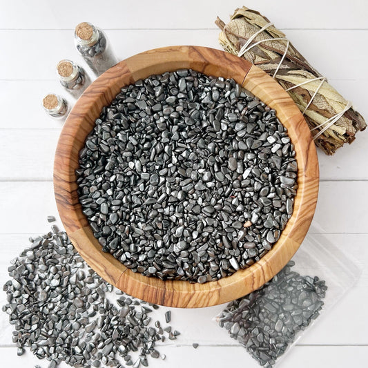 A wooden bowl filled with small, smooth black Hematite Crystal Chips sits on a white surface. Surrounding the bowl are three small glass jars with cork tops containing Hematite Crystal Chips, a bundle of dried sage wrapped in string, and a scattered pile of stones next to a plastic bag. Ideal for handmade crafts and grounding protection.