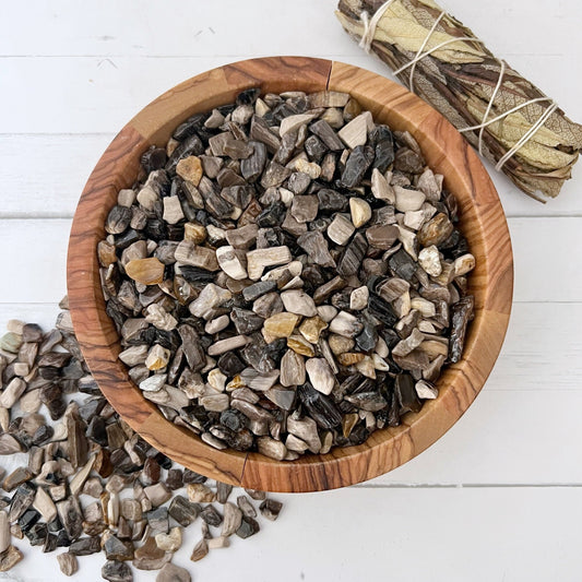 A wooden bowl is filled with various small polished stones in shades of brown, beige, white, and gray. Among them are Petrified Wood Crystal Chips known for grounding energies. Some Petrified Wood Crystal Chips are scattered beside the bowl on a white wooden surface, while a bundle of dried sage, wrapped in string, lies next to it.