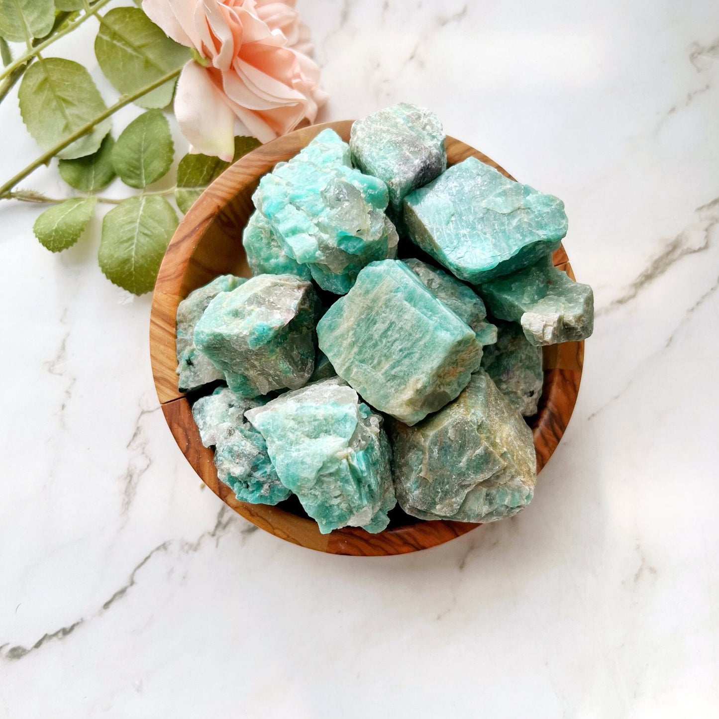 A wooden bowl filled with Raw Amazonite Stones is placed on a white marble surface. There is a pink rose with green leaves resting next to the bowl, adding a touch of elegance to the serene setting. The rough stones create an enchanting contrast against the smooth marble.