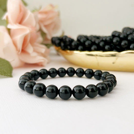 A polished 8mm Black Obsidian Beaded Bracelet is displayed on a white surface, with a bunch of pink roses in the background. Additional black beads, known for their protective qualities, are scattered on a gold-colored tray behind the bracelet, contributing to the elegant and serene composition.