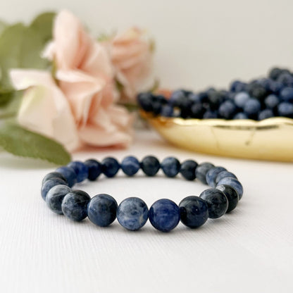 A 8mm Sodalite Beaded Bracelet lies on a white surface. In the background, there are soft pink roses and a bowl filled with blueberries, both slightly out of focus. The setting gives a serene and elegant feel, emphasizing the bracelet's metaphysical properties.