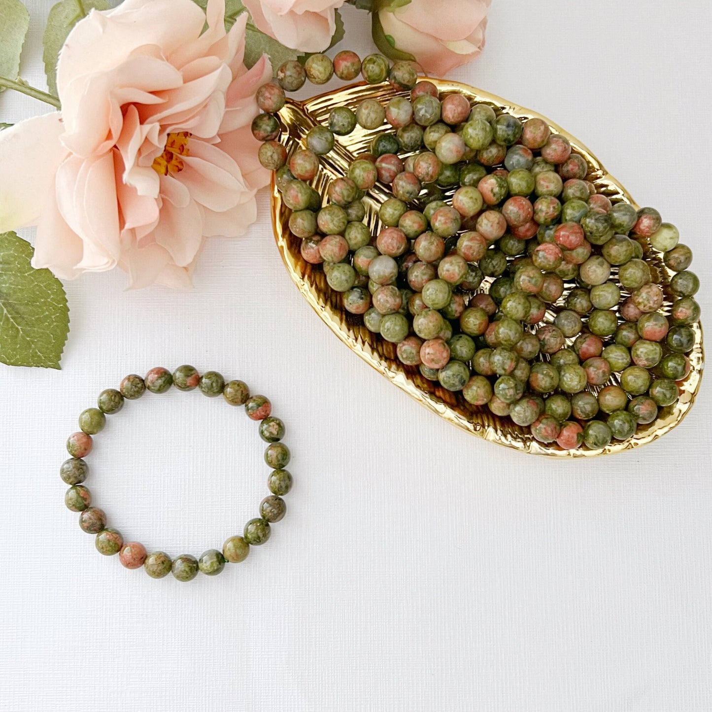 An 8mm Unakite Beaded Bracelet made of green and pink stones is displayed on a white surface. In the background, there is a blurred image of pink flowers and a golden dish filled with similar stones. The overall setting, emphasizing emotional healing, conveys a natural and elegant aesthetic.