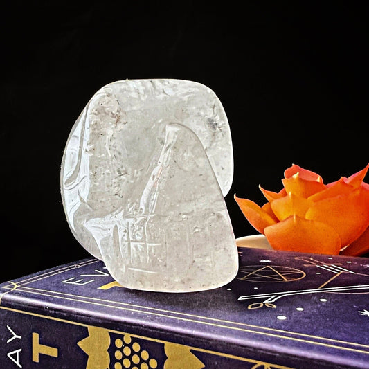 A smooth, translucent 2" Clear Quartz Crystal Skull sits on a purple book adorned with golden astrological symbols. An orange flower is placed beside the sculpture, providing a vivid contrast against the dark background, infusing the scene with serene Reiki energy.