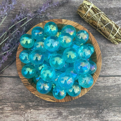 A wooden bowl filled with shiny, iridescent Mini Aqua Aura Quartz Crystal Spheres sits on a gray wooden surface. To the left, there is a bundle of lavender, and to the right, a neatly tied bundle of sage, adding a rustic and natural touch to the scene. The spheres radiate a healing aura reminiscent of Aqua Aura Quartz.