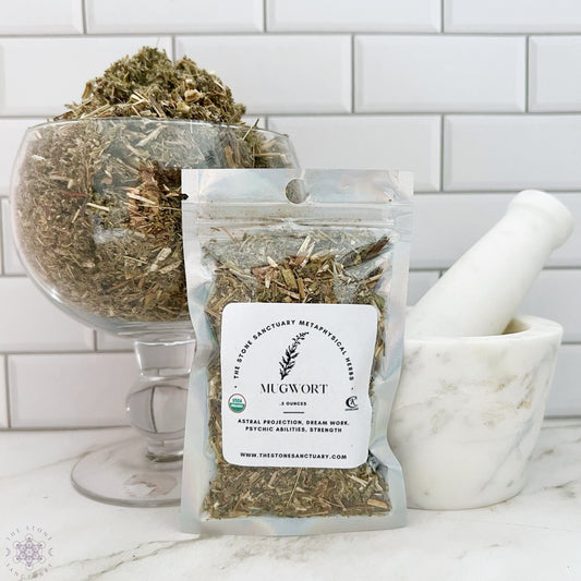 A small pouch labeled "Mugwort Herb" filled with dried herbs is placed in front of a transparent glass container brimming with more dried herbs. Next to it, there is a white mortar and pestle, all set against a white tiled background—perfect for divination practices.