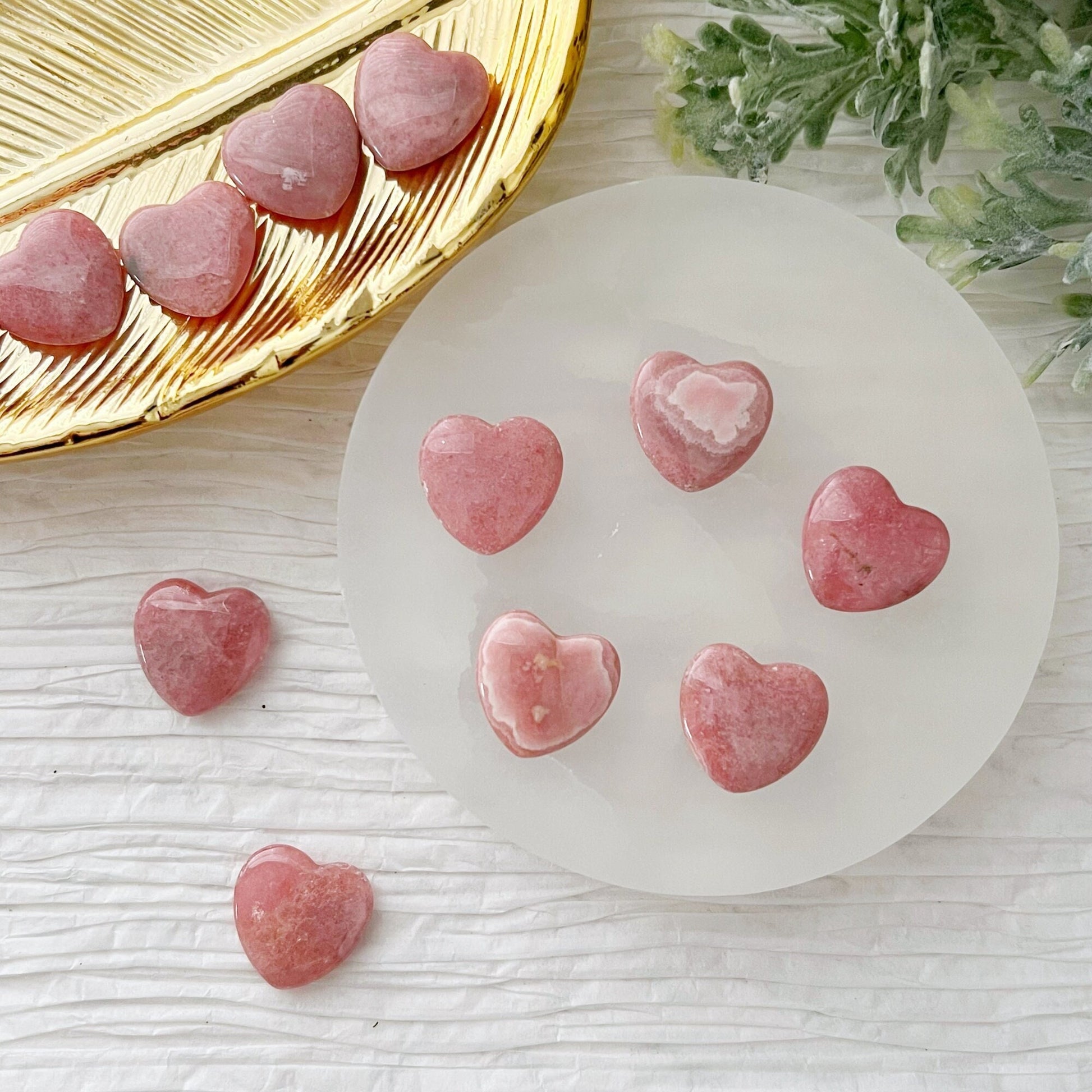 20mm Mini Rhodochrosite Hearts, known as the stone of love, are elegantly arranged on a circular, translucent white plate. Additional stones rest on a golden leaf-shaped dish and a textured white surface. Green foliage is partially visible in the upper right corner of the image, enhancing the heart chakra energy.