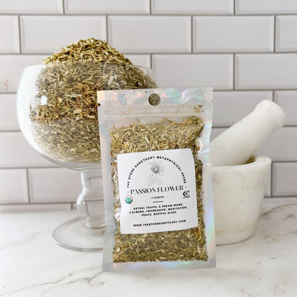 A glass bowl filled with dried herbs is in the background with some herbs in a dish. In the foreground, a clear plastic bag labeled "Passion Flower" containing more dried herbs stands upright. A white mortar and pestle are on the right. All items are on a white surface.