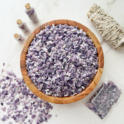 A wooden bowl filled with small Lepidolite Crystal Chips sits on a white surface, with additional Lepidolite Crystal Chips scattered nearby. Short glass bottles filled with the same crystals and a bound bundle of dried sage, perfect for crafting or metaphysical uses, are also placed on the surface.