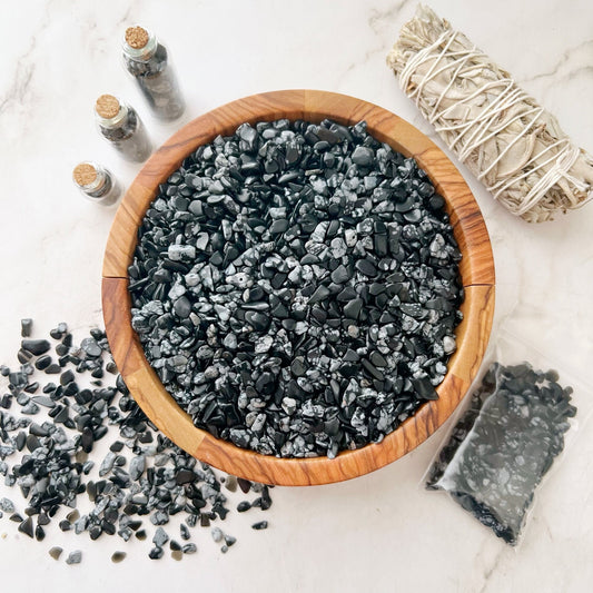 Image of a wooden bowl filled with polished black and frosty white Snowflake Obsidian Chips on a white surface. A pile of crystal chips rests outside the bowl on the left, with a few in small glass jars. To the right, there's a bundle of sage and a sealed plastic bag of stones, perfect for meditation sessions.