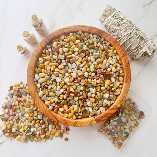 A wooden bowl filled with multicolored polished stones is placed on a white surface. To the left, small piles of Crazy Lace Agate Crystal Chips and several small jars brim with similar stones. To the right, a bundle of sage and a small plastic bag containing crystal chips are visible, radiating natural energy.