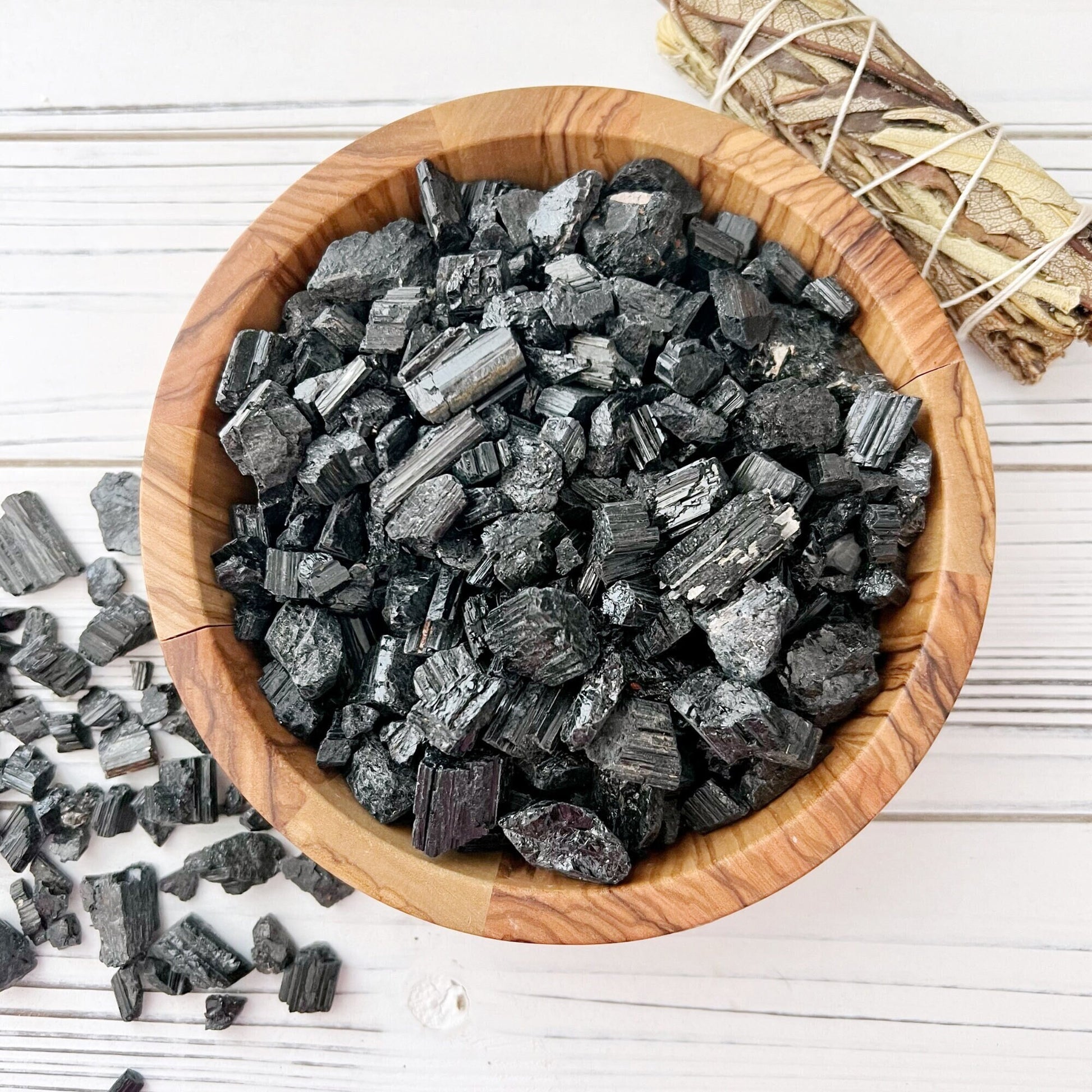 A wooden bowl filled with Rough Black Tourmaline Chips, known for their protective energy, sits on a light wooden surface. Some tourmaline chips are scattered near the bowl. Beside the bowl, there is a bundle of sage wrapped with twine.