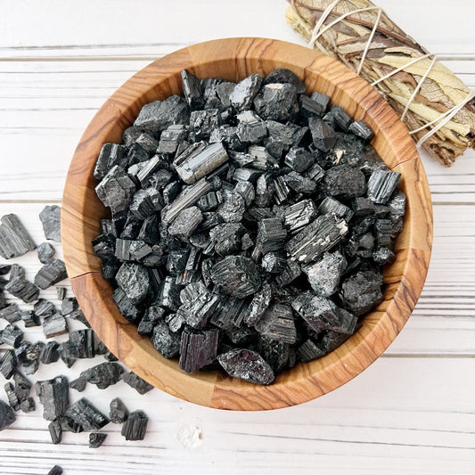 A wooden bowl filled with Rough Black Tourmaline Chips, known for their protective energy, sits on a light wooden surface. Some tourmaline chips are scattered near the bowl. Beside the bowl, there is a bundle of sage wrapped with twine.